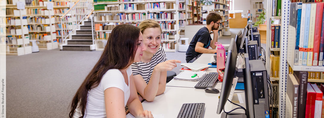 Zwei Personen sitzen nebneinander in der Unibibliothek, beide schauen auf einen Bildschirm